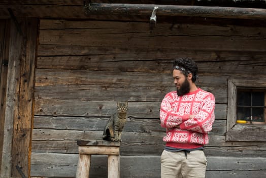 portrait of young hipster man with beard and cat in front of wooden house