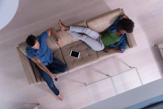 Young multiethnic couple relaxes on the sofa in the luxury living room, using a tablet and remote control top view
