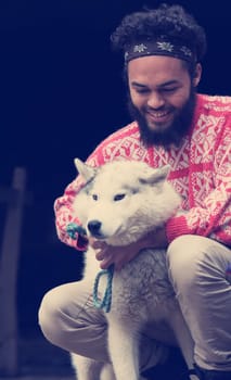 handsome young hipster man playing with white husky dog in front of old vintage retro wooden house