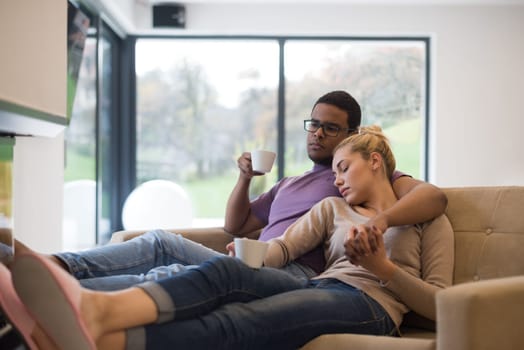 Young romantic multiethnic couple sitting on sofa in front of fireplace at home, looking at each other, talking and drinking coffee
