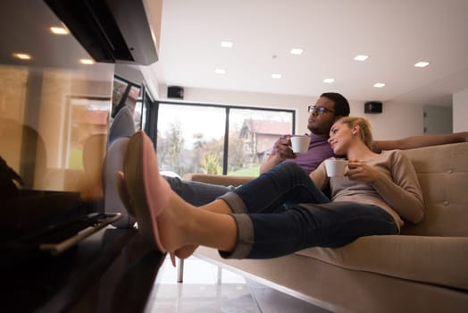 Young romantic multiethnic couple sitting on sofa in front of fireplace at home, looking at each other, talking and drinking coffee