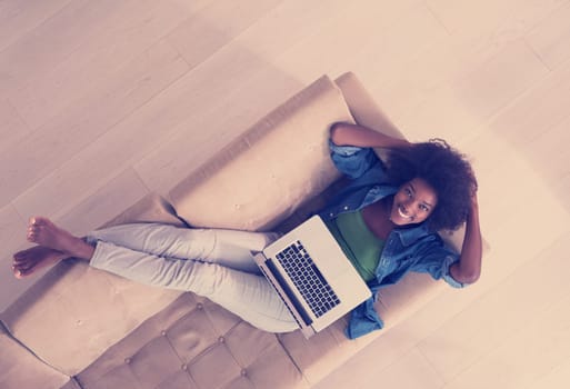 Smiling young African American woman using laptop while relaxing on sofa at luxury home top view