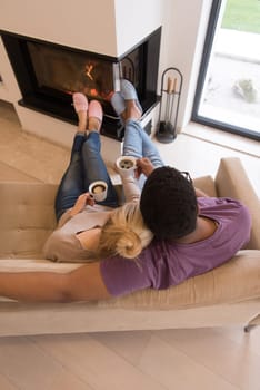 Young romantic multiethnic couple sitting on sofa in front of fireplace at home, looking at each other, talking and drinking coffee