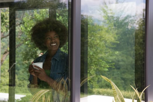 Beautiful young african american woman drinking coffee and looking through a window in her luxury home