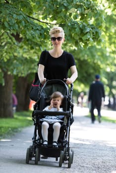 beautiful young mother with blond hair and sunglasses pushed her baby daughter in a stroller on a summer day