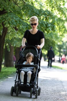 beautiful young mother with blond hair and sunglasses pushed her baby daughter in a stroller on a summer day