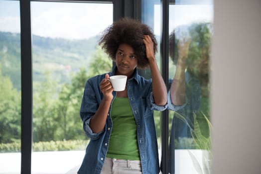 Beautiful young african american woman drinking coffee and looking through a window in her luxury home