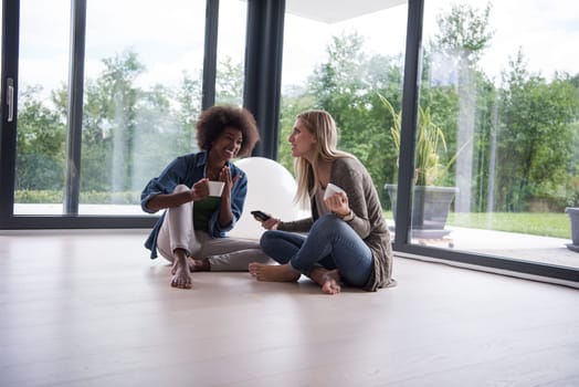 Two young laughing multiethnic women sit on the floor near the window and enjoy while drinking coffee using smartphone in luxury home