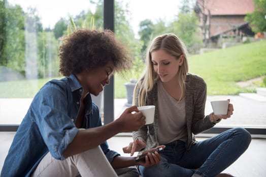 Two young laughing multiethnic women sit on the floor near the window and enjoy while drinking coffee using smartphone in luxury home