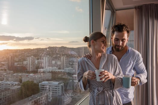romantic happy young couple enjoying evening coffee and beautiful sunset landscape of the city while standing by the window