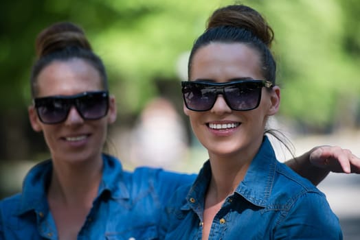 Young beautiful twin sister with sunglasses in identical wardrobe posing in a park on a sunny summer day