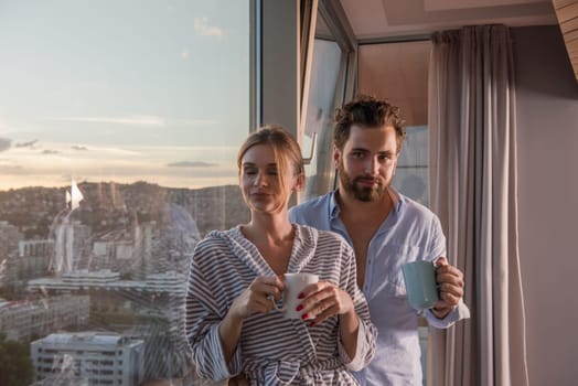 romantic happy young couple enjoying evening coffee and beautiful sunset landscape of the city while standing by the window