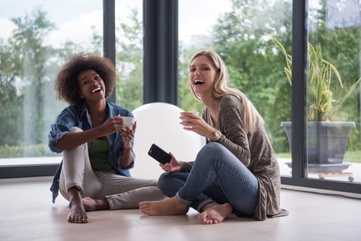 Two young laughing multiethnic women sit on the floor near the window and enjoy while drinking coffee using smartphone in luxury home
