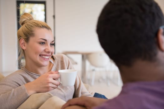 Young romantic multiethnic couple sitting on sofa in front of fireplace at home, looking at each other, talking and drinking coffee