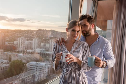 romantic happy young couple enjoying evening coffee and beautiful sunset landscape of the city while standing by the window