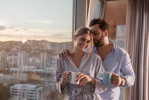 romantic happy young couple enjoying evening coffee and beautiful sunset landscape of the city while standing by the window