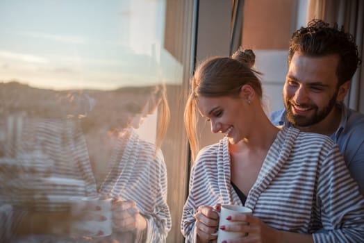 romantic happy young couple enjoying evening coffee and beautiful sunset landscape of the city while standing by the window