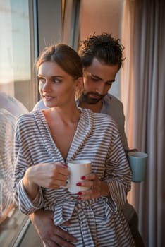 romantic happy young couple enjoying evening coffee and beautiful sunset landscape of the city while standing by the window