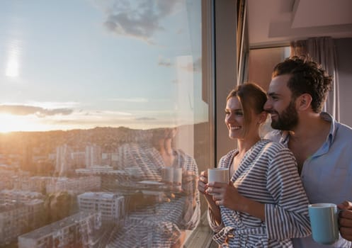 romantic happy young couple enjoying evening coffee and beautiful sunset landscape of the city while standing by the window