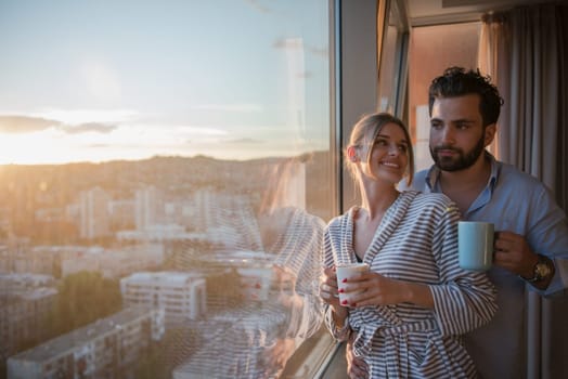 romantic happy young couple enjoying evening coffee and beautiful sunset landscape of the city while standing by the window