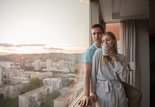 romantic happy young couple enjoying evening coffee and beautiful sunset landscape of the city while standing by the window