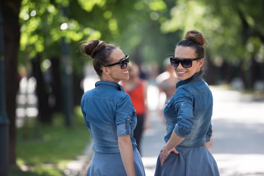 Young beautiful twin sister with sunglasses in identical wardrobe posing in a park on a sunny summer day