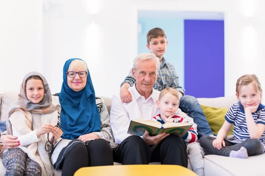 modern muslim family grandparents with grandchildren reading Quran and praying together on the sofa before iftar dinner during a ramadan feast at home