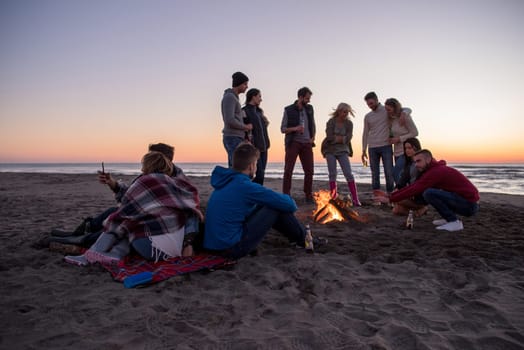 Happy Carefree Young Friends Having Fun And Drinking Beer By Bonefire On The Beach As The Sun Begins To Set