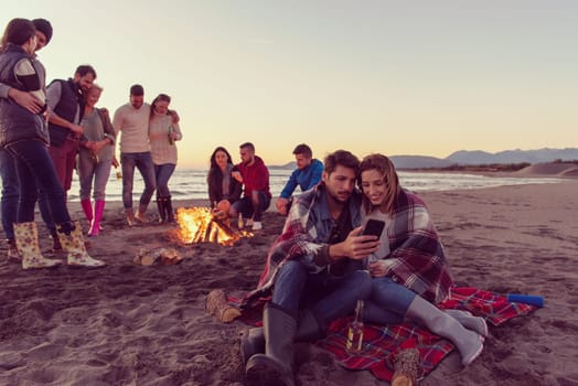 Couple using cell phone during autumn beach party with friends drinking beer and having fun