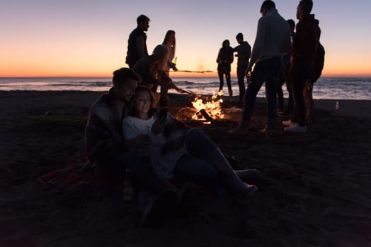 Couple using cell phone during autumn beach party with friends drinking beer and having fun