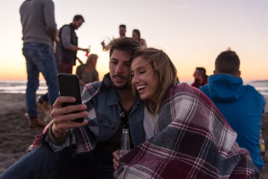 Couple using cell phone during autumn beach party with friends drinking beer and having fun