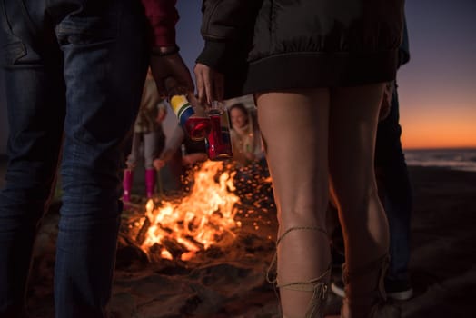 Happy Carefree Young Friends Having Fun And Drinking Beer By Bonefire On The Beach As The Sun Begins To Set
