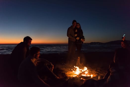Happy Carefree Young Friends Having Fun And Drinking Beer By Bonefire On The Beach As The Sun Begins To Set