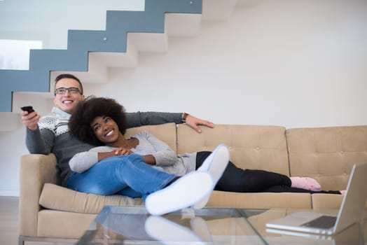 Young multiethnic couple relaxing on the sofa in the luxury living room, using a laptop and remote control