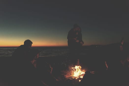 Happy Carefree Young Friends Having Fun And Drinking Beer By Bonefire On The Beach As The Sun Begins To Set