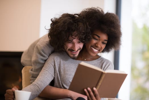 Young beautiful multiethnic couple hugging in front of fireplace at home when reading a book at autumn day