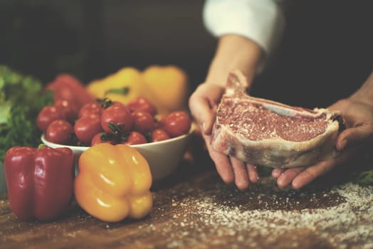 Master Chef holding juicy slice of raw steak with vegetables around on a wooden table