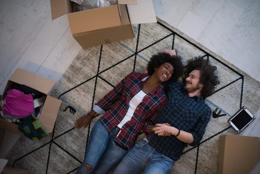 Top view of attractive young multiethnic couple moving, holding hands, looking at camera and smiling while lying among cardboard boxes