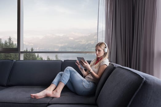 Young beautiful girl enjoying music through headphones, laying on sofa near the window at home