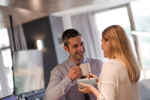 A young couple is preparing for the job and using a laptop. The man drinks coffee while the woman eats breakfast at luxury home together, looking at screen, smiling.