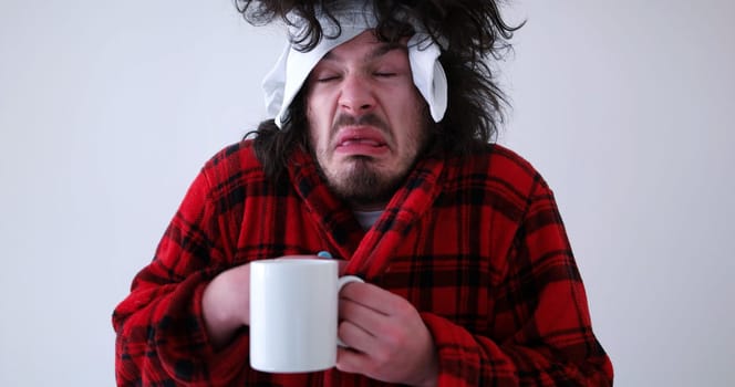 young Man with flu and fever wrapped holding cup of healing tea isolated over white
