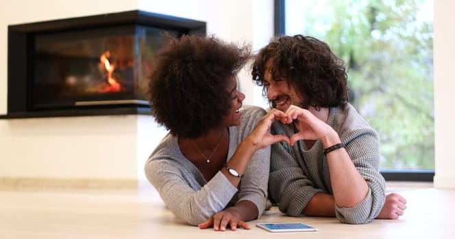 beautiful young multiethnic couple with tablet computer showing a heart with their hands on the floor in front of fireplace at autumn day