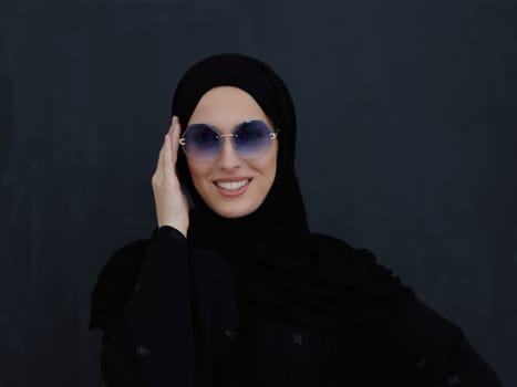 Young muslim in traditional clothes or abaya and sunglasses posing in front of black chalkboard. Arab woman representing modern arabic lifestyle, islamic fashion and Ramadan kareem concept