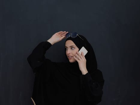 Young muslim businesswoman in traditional clothes or abaya talking on the smartphone. Arab woman in front of black chalkboard and representing techology, islamic fashion and Ramadan kareem concept