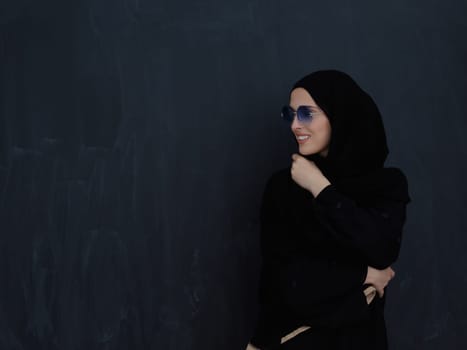 Young muslim in traditional clothes or abaya and sunglasses posing in front of black chalkboard. Arab woman representing modern arabic lifestyle, islamic fashion and Ramadan kareem concept