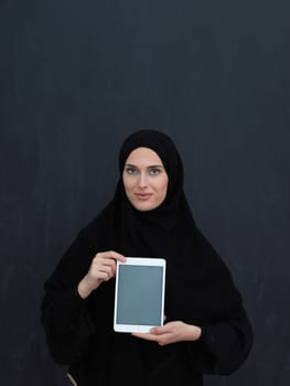 Young Arab businesswoman in traditional clothes or abaya and glasses showing tablet computer display in front of black chalkboard representing islam fashion and technology