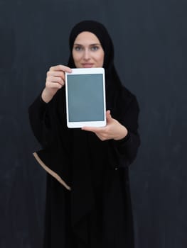 Young Arab businesswoman in traditional clothes or abaya and glasses showing tablet computer display in front of black chalkboard representing islam fashion and technology