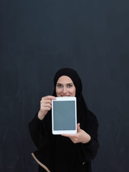 Young Arab businesswoman in traditional clothes or abaya and glasses showing tablet computer display in front of black chalkboard representing islam fashion and technology