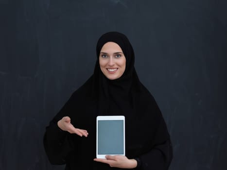 Young Arab businesswoman in traditional clothes or abaya and glasses showing tablet computer display in front of black chalkboard representing islam fashion and technology