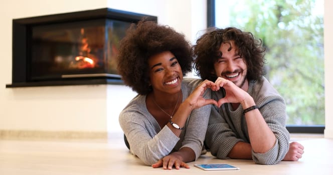beautiful young multiethnic couple with tablet computer showing a heart with their hands on the floor in front of fireplace at autumn day
